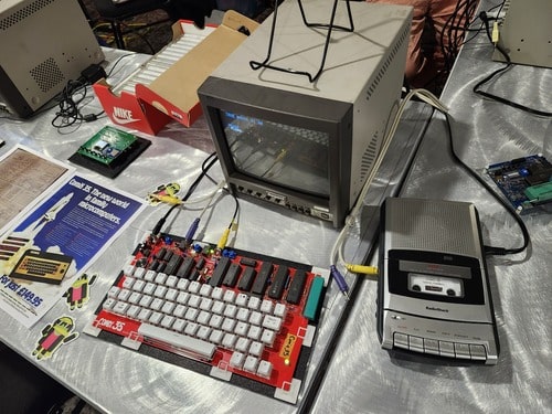 Photo of a COMX-35 computer at the ATG Expo. The COMX-35 computer has no case, just the bare motherboard and keyboard. A tape recorder is connected for loading software, and a security camera monitor is connected as a display. Surrounding the computer are copies of ads for the COMX-35, and a shoebox full of cassette tapes featuring different programs for the computer.