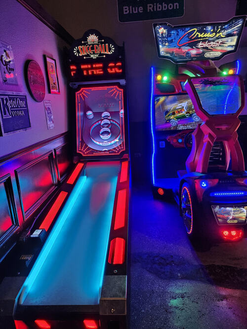 Photo of games at Crickets Draft House & Grill in Waco, Texas. On the left is a skee-ball game with dramatic red-and-blue lighting. On the right side is a sit-down Cruis’n Blast game. Hanging from the left wall are signs for brands like Ketel One vodka and Pabst Blue Ribbon beer.
