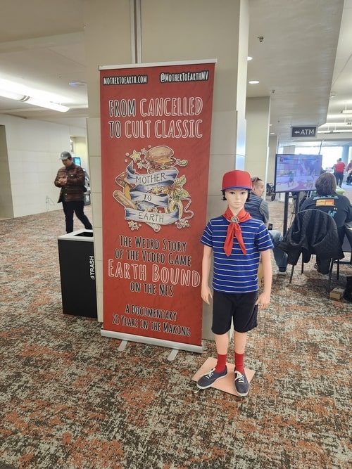 Photo from ATG Expo showing a banner for the documentary Mother to Earth, next to mannequin of the character Ninten from the game Mother 1. Ninten is a boy with black hair, a white-and-blue striped shirt, a red ball cap (red with a blue brim), black shorts, red socks, and sneakers. 