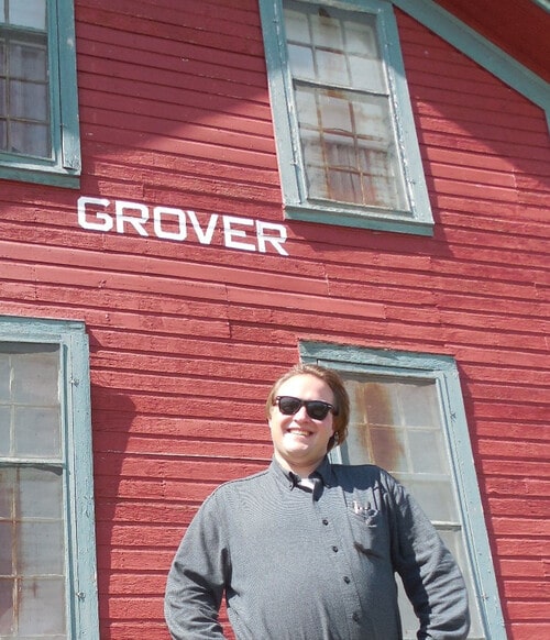 Photograph of Nicholas Bernhard in a green lumberjack shirt and sunglasses standing in front of the Grover Depot Museum, a red two-story wooden building with “Grover” stenciled in white lettering about halfway up. Photo by Nicholas Bernhard, licensed under Creative Commons Attribution 4.0.