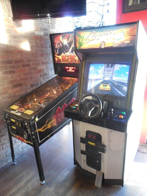 Left to right, an AC/DC pinball machine, and an upright Cruis’n USA cabinet in a restaurant designed to resemble old-time NYC pizzerias, with brick walls on the size. 