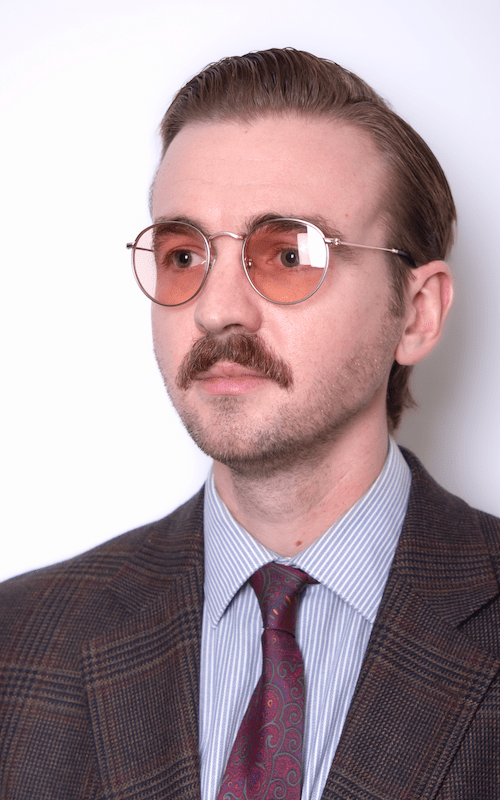 Photograph of author Sefton Eisenhart. He is wearing a suit and tie, with a mustache and orange-tinted glasses. He is standing in front of a white background.